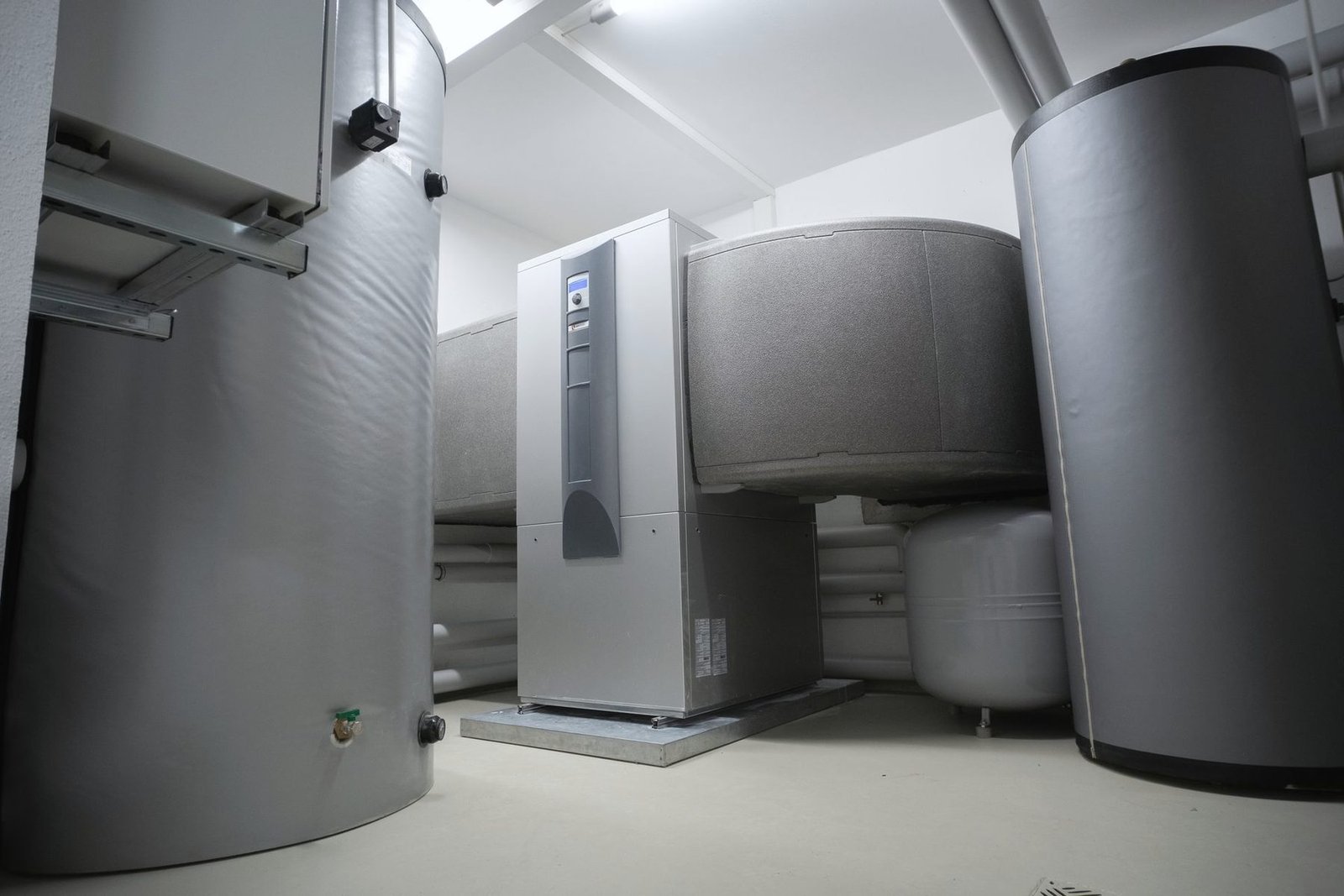 A clean mechanical room with insulated storage tanks and a grey heating unit on a concrete floor
