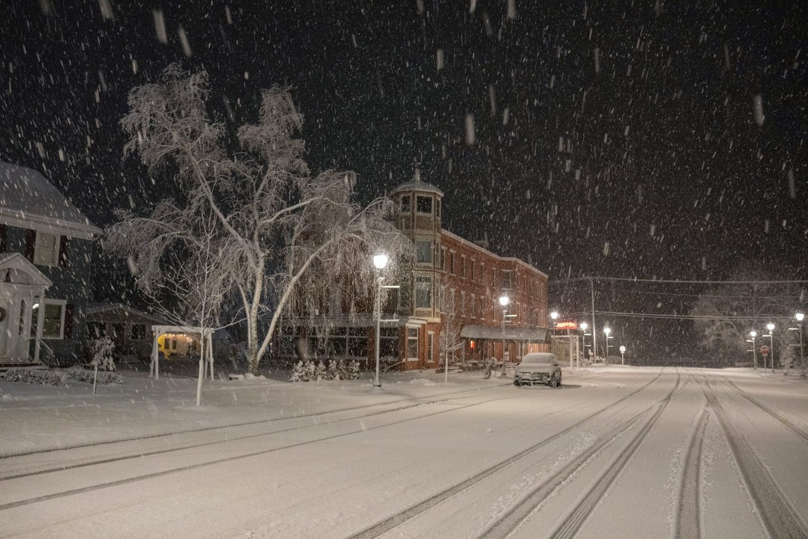 A quiet small-town street at night under falling snow, lit by street lamps